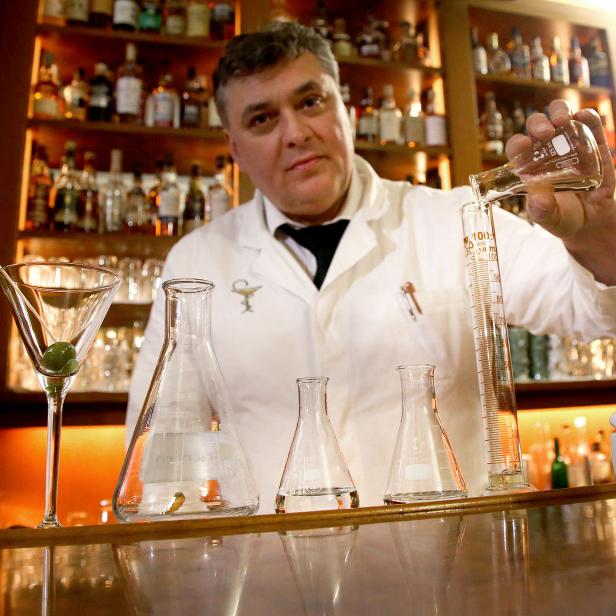 Ein Barkeeper im Laborkittel mixt einen Drink mit Messzylinder und Kolben vor einer Bar.
