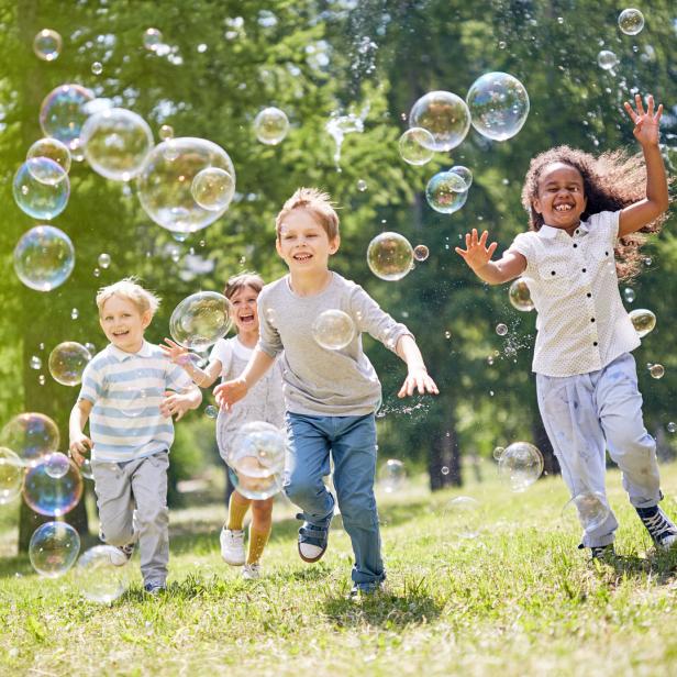 Vier fröhliche Kinder rennen durch eine Wiese voller Seifenblasen.