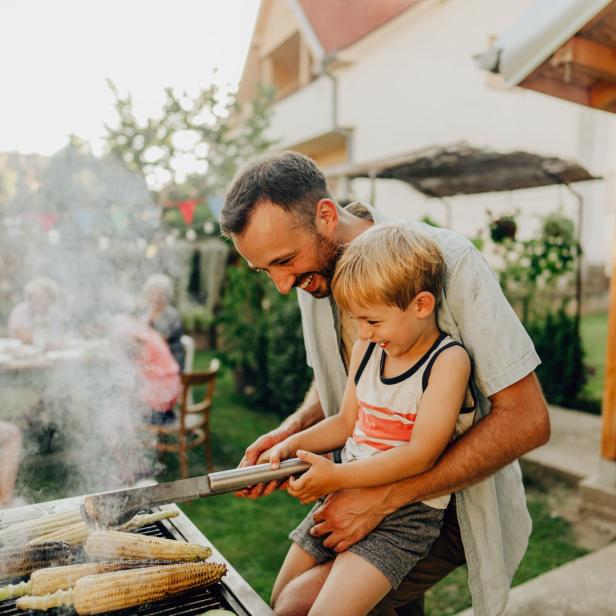 Ein Vater grillt mit seinem kleinen Sohn Mais und Zucchini im Garten.