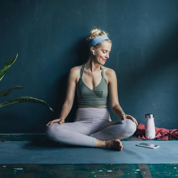Eine Frau sitzt mit Kopfhörern auf einer Yogamatte und meditiert.