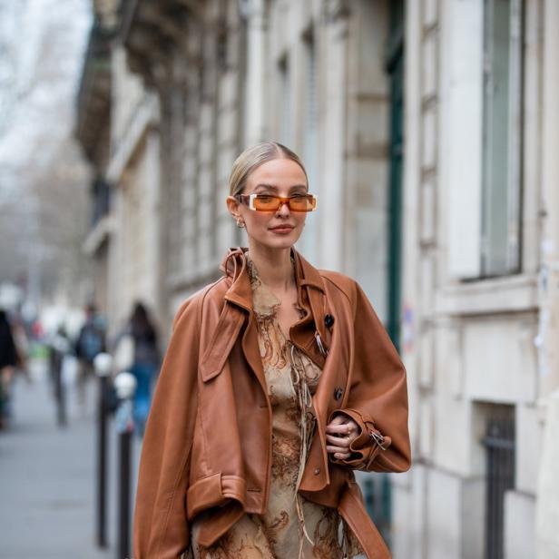Eine Frau mit orangefarbener Sonnenbrille, brauner Jacke und passender Handtasche geht die Straße entlang.