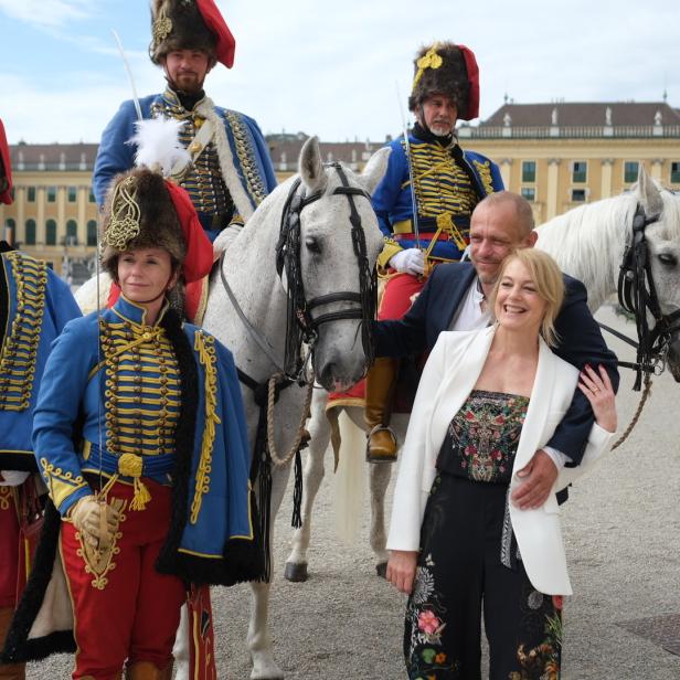 Eine Frau posiert mit Männern in historischen Husarenuniformen und weißen Pferden vor Schloss Schönbrunn.