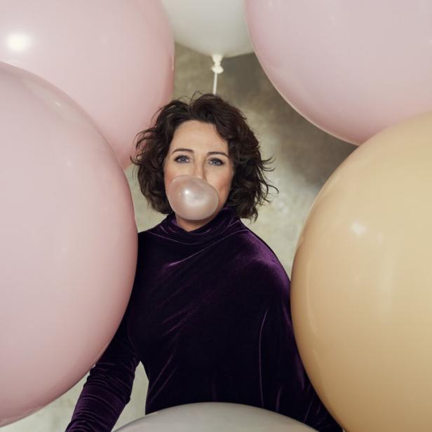 Eine Frau mit dunklen Haaren macht eine Kaugummiblase, umgeben von pastellfarbenen Ballons.