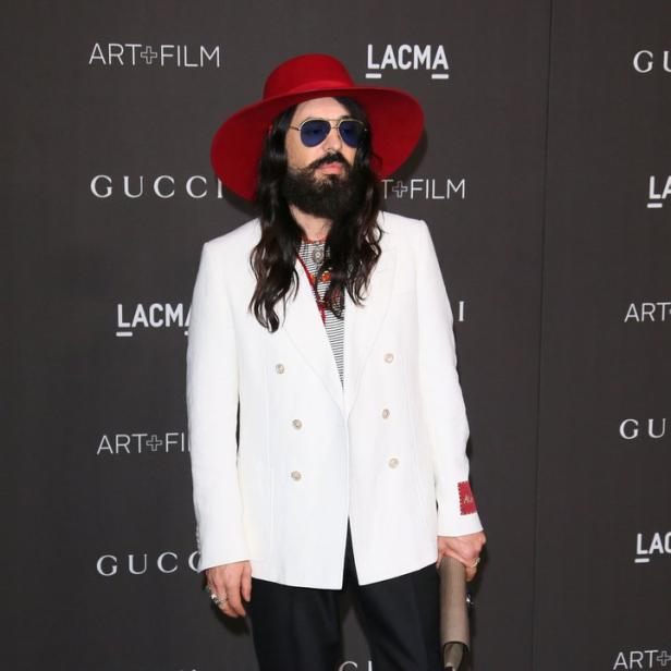Ein Mann mit Bart und rotem Hut posiert vor einem Gucci-Logo.