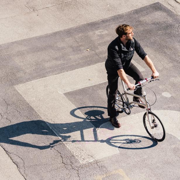 Ein Mann mit Bart fährt mit einem silbernen Fahrrad über eine graue Straße.