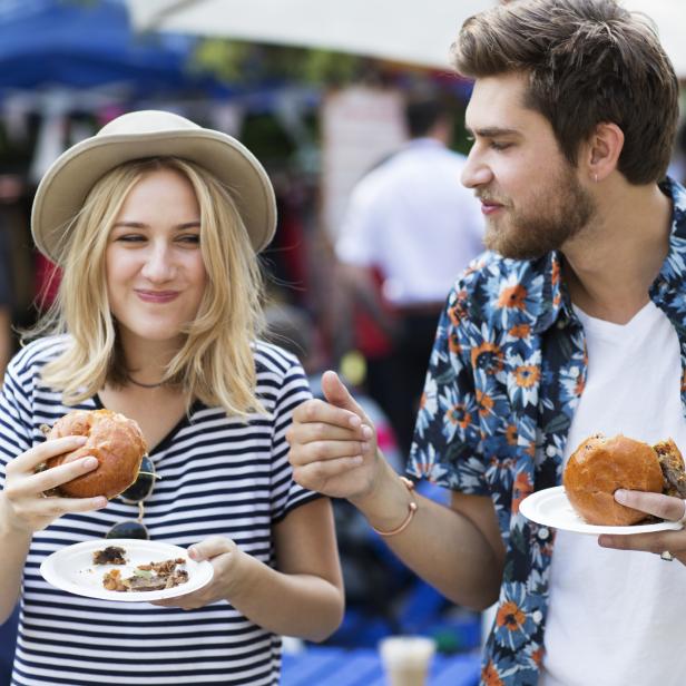 Eine Frau mit Hut und ein Mann essen Burger auf einem Markt.