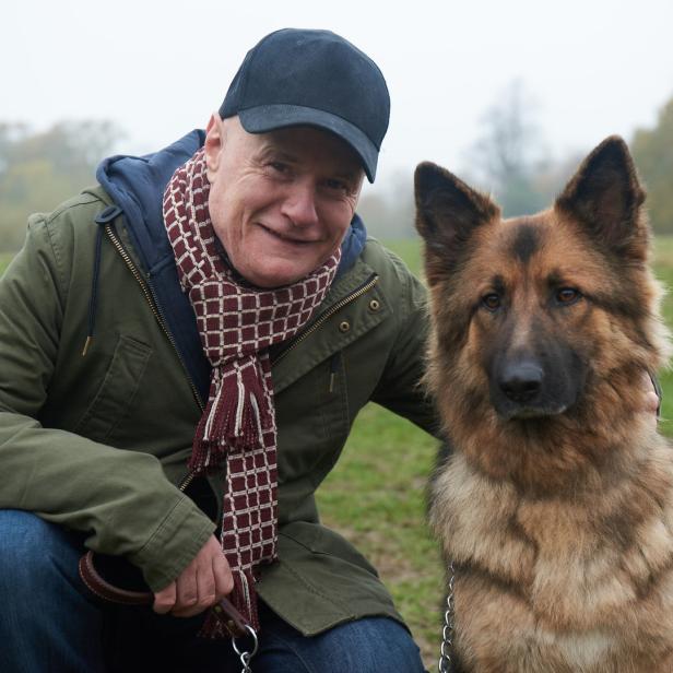 Ein Mann kniet neben einem Deutschen Schäferhund auf einer Wiese.