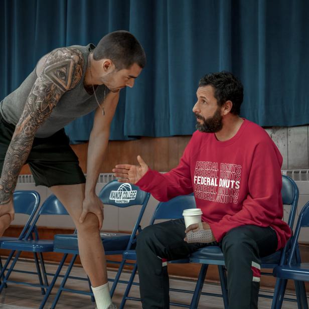 Adam Sandler sitzt mit einem Kaffee in der Hand und spricht mit einem tätowierten Mann.