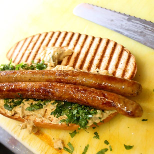 Ein Sandwich mit Würstchen, Kräutern und Soße auf einem gelben Schneidebrett.