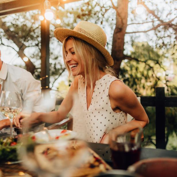 Eine lachende Frau mit Hut sitzt mit einem Mann bei einem Abendessen im Freien.
