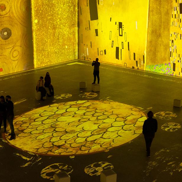 Besucher in einer Ausstellung, in der Werke von Gustav Klimt auf Wände und Boden projiziert werden.