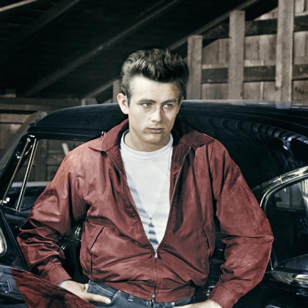 James Dean posiert an einem Auto in roter Jacke und Jeans.