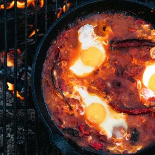 Shakshuka wird in einer gusseisernen Pfanne über einem Lagerfeuer zubereitet.