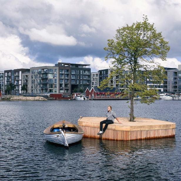 Ein Mann sitzt auf einer schwimmenden Plattform mit Baum, ein Boot liegt daneben.