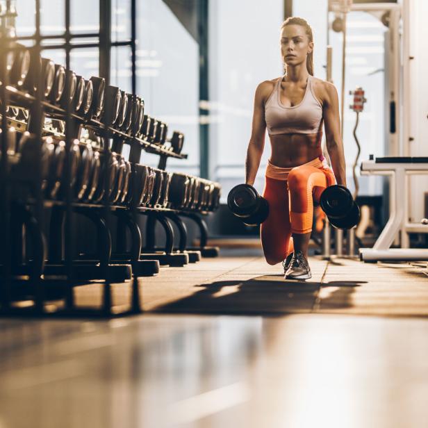 Female athlete exercising with dumbbells in a lunge position at gym.