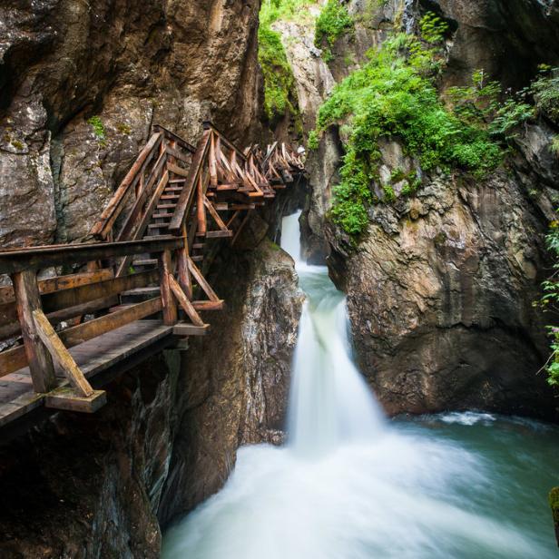 Ein hölzerner Steg führt über einen tosenden Wasserfall in einer engen Schlucht.