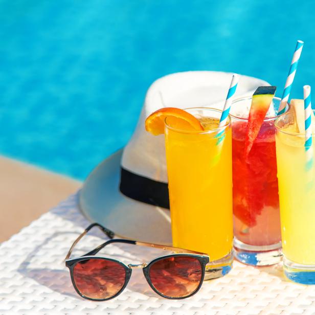 Drei Cocktails mit Strohhalmen stehen neben einem Hut und einer Sonnenbrille am Pool.