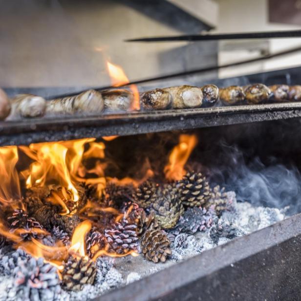 Würste und Fleisch werden über einem Feuer aus Kiefernzapfen gegrillt.