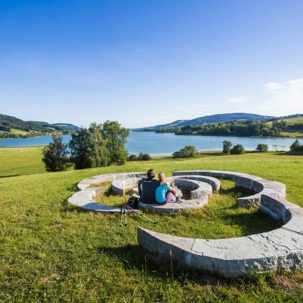 Ein Paar sitzt auf einer spiralförmigen Steinbank mit Blick auf einen See.