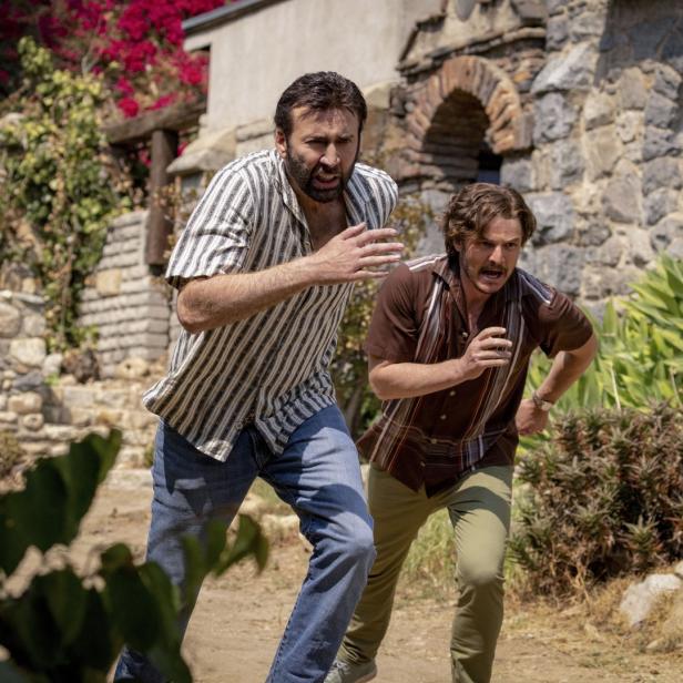Nicolas Cage und Pedro Pascal rennen in „Massive Talent“ nebeneinander durch eine üppige Landschaft.