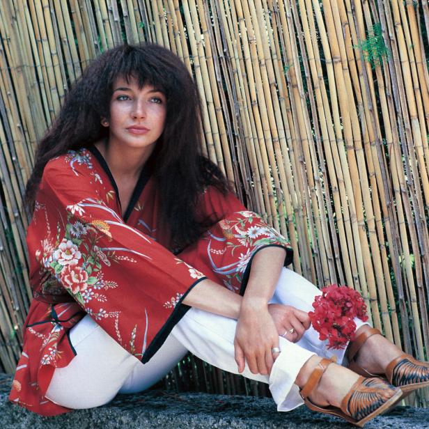 Kate Bush sitzt vor einem Bambushintergrund und hält rote Blumen.