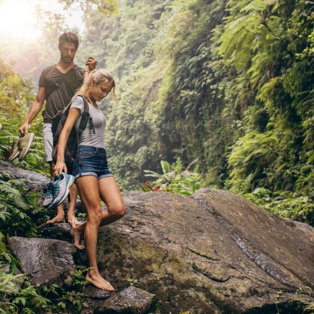 Ein Paar wandert barfuß durch einen üppigen Wald und trägt die Schuhe in der Hand.