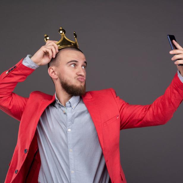 Ein Mann mit Bart und rotem Sakko macht ein Selfie mit einer goldenen Krone auf dem Kopf.