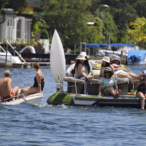 Eine Gruppe von Menschen entspannt sich auf einem Floß mit Palme, während ein Segelboot vorbeifährt.