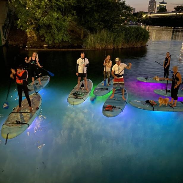 Eine Gruppe von Menschen steht auf beleuchteten Stand-Up-Paddleboards auf einem Fluss bei Dämmerung.