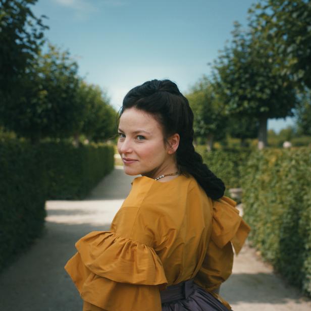 Eine Frau in historischer Kleidung blickt in einem Garten über ihre Schulter.