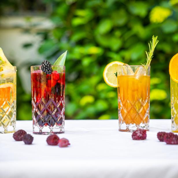 Vier Gläser mit bunten Cocktails, garniert mit Früchten und Kräutern, stehen auf einem Tisch.