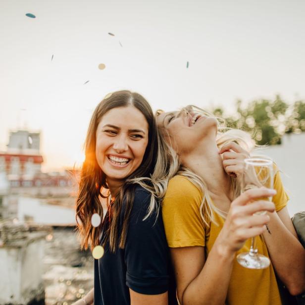 Drei junge Frauen feiern lachend mit Konfetti und Sekt auf einer Dachterrasse.