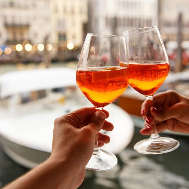 Zwei Personen stoßen mit Aperol Spritz vor einer venezianischen Kulisse an.