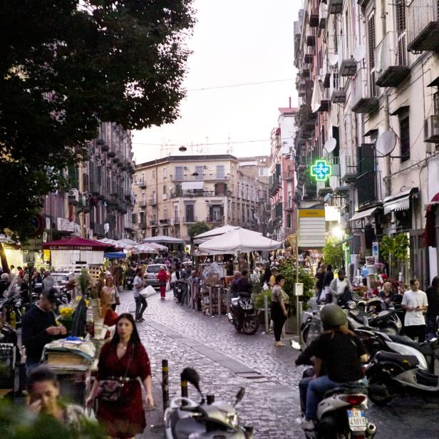 Eine belebte Straße in Neapel mit Menschen, Marktständen und geparkten Motorrollern.