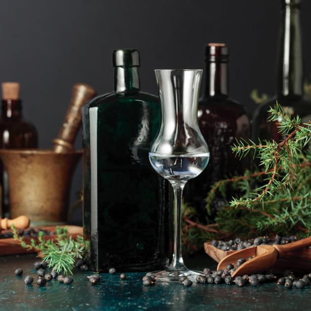 Ein Glas klarer Schnaps steht neben Wacholderbeeren und Flaschen.