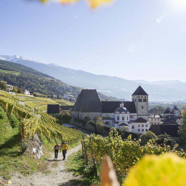 Zwei Personen wandern durch die Weinberge mit Blick auf ein Kloster und die Berge.