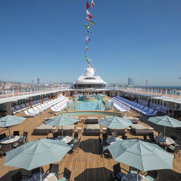 Blick von oben auf das Deck eines Kreuzfahrtschiffes mit Pool, Liegestühlen und Tischen mit Sonnenschirmen.