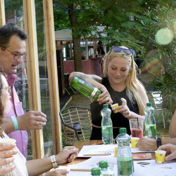Eine Gruppe von Menschen sitzt an einem Tisch, während eine Frau Getränke aus einer grünen Flasche einschenkt.