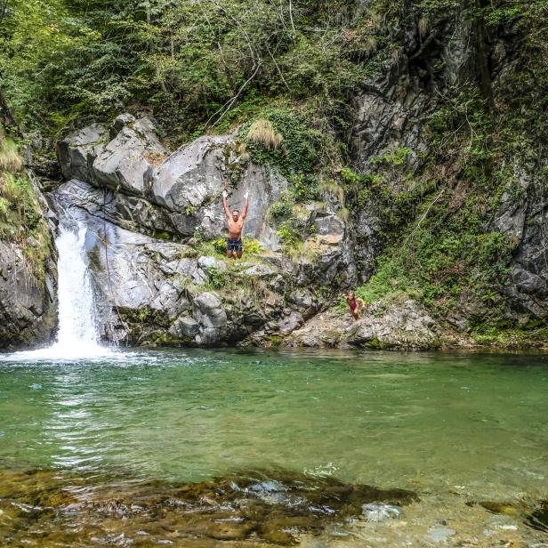 Ein Mann steht mit erhobenen Armen auf einem Felsen neben einem Wasserfall, während ein Kind ins Wasser springt.