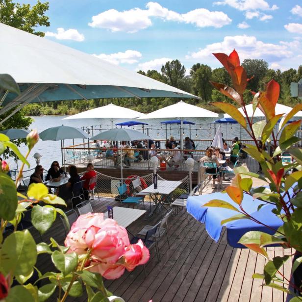Ein Restaurant am See mit Gästen unter Sonnenschirmen auf einer Terrasse.