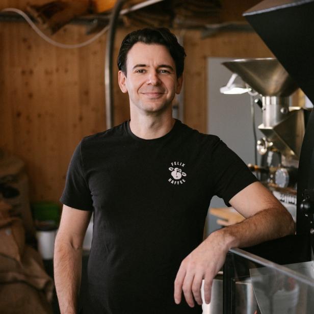 Ein Mann mit einem T-Shirt der Marke „Felix Kaffee“ steht neben einer Kaffeeröstmaschine.