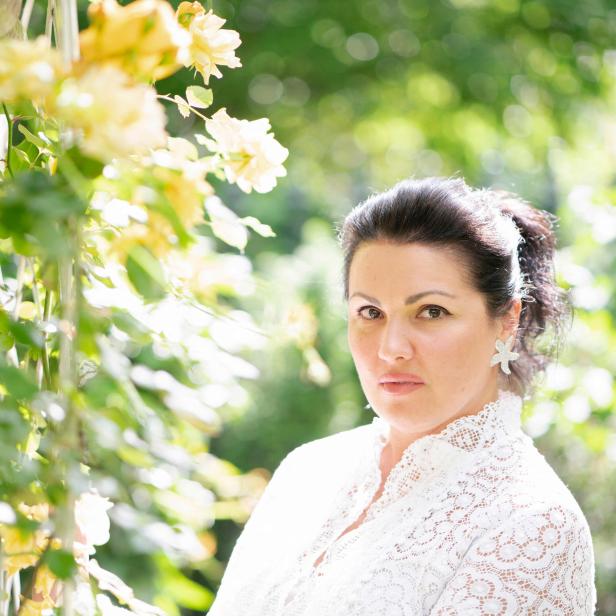 Eine Frau in einem weißen Spitzenoberteil steht vor gelben Rosen.
