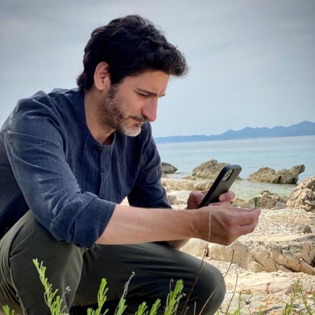 Ein Mann hockt am Strand und betrachtet eine Pflanze, während er sein Handy hält.
