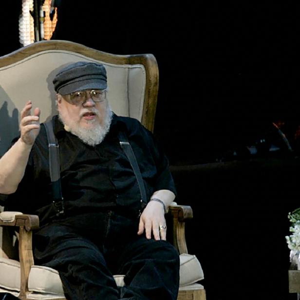 George R.R. Martin sitzt in einem Sessel und gestikuliert während einer Veranstaltung.