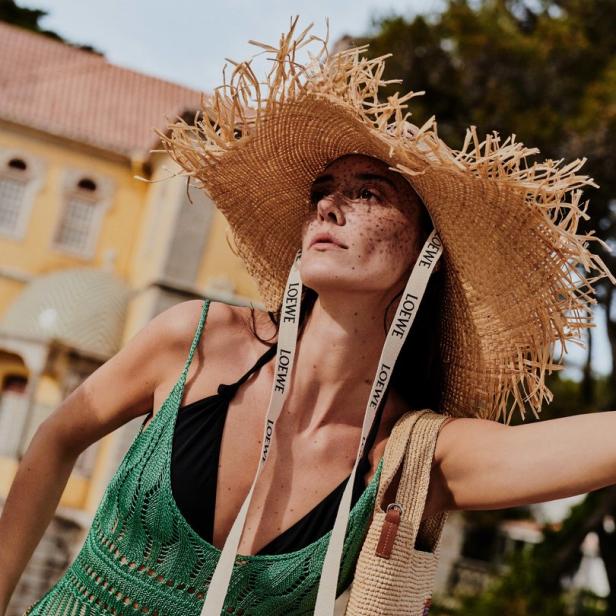 Eine Frau mit Strohhut, grünem Kleid und einer Tasche der Marke Loewe vor einem Gebäude.