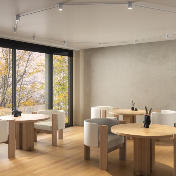 Ein heller Raum mit Holzboden, Tischen und Stühlen vor einem großen Fenster mit Herbstbäumen.