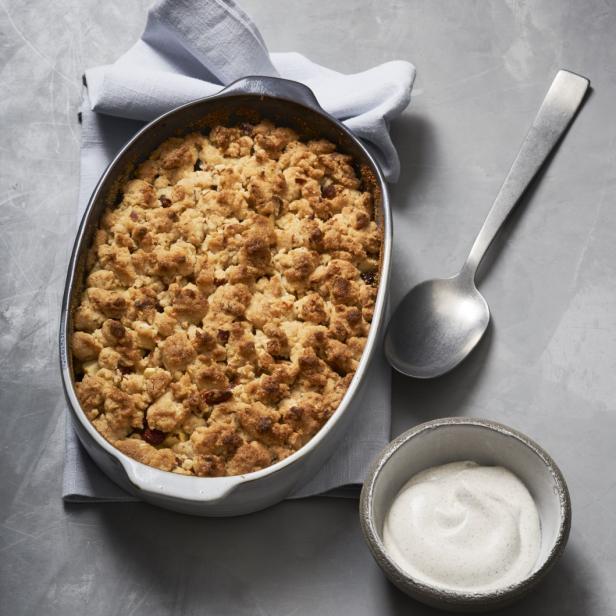 Ein Apfel-Crumble in einer Form mit einem Löffel und einer Schale Creme auf einem grauen Hintergrund.