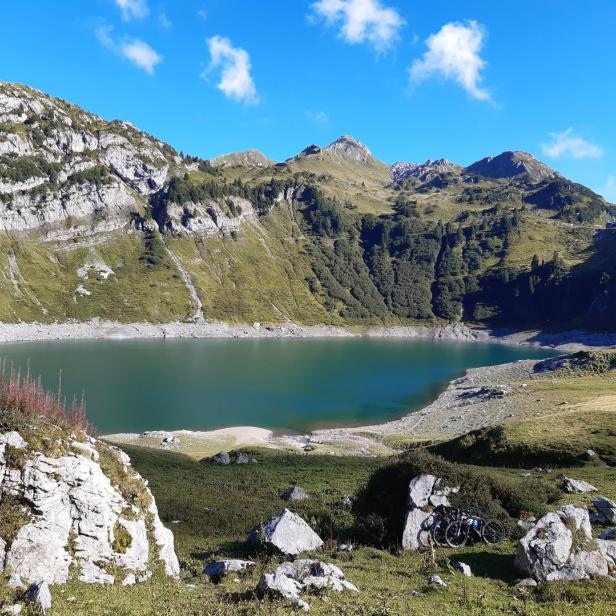 Ein Bergsee liegt eingebettet in eine grüne Berglandschaft unter blauem Himmel. Im Vordergrund stehen zwei Fahrräder.