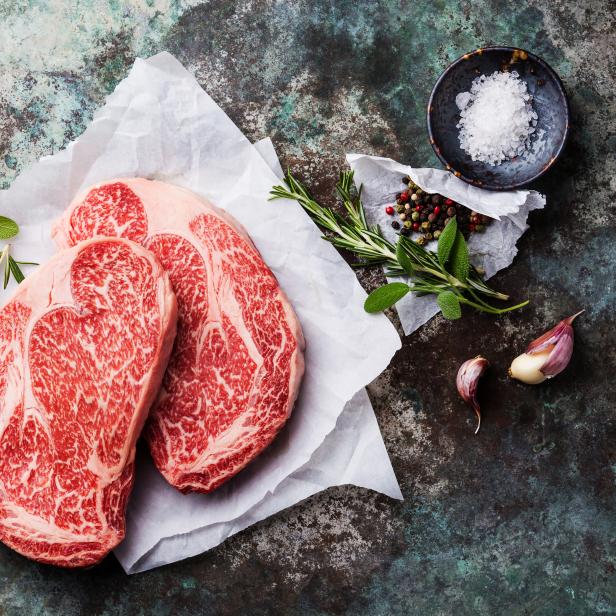 Zwei rohe Ribeye-Steaks mit Marmorierung, zusammen mit Gewürzen wie Rosmarin, Salbei, Pfeffer und Knoblauch.