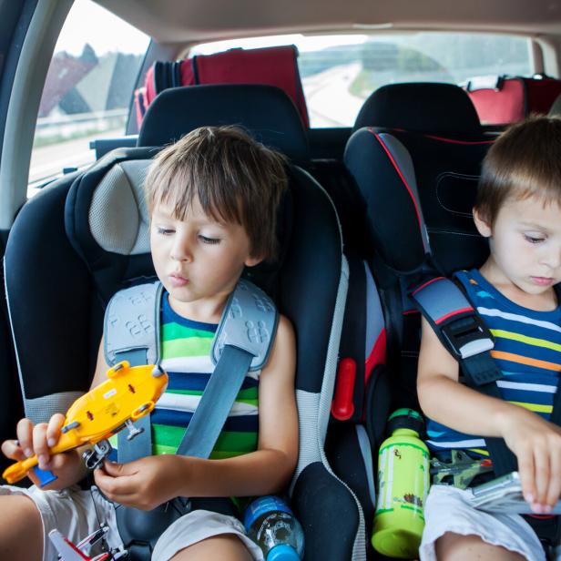 Zwei kleine Jungen in gestreiften T-Shirts sitzen in Kindersitzen auf der Rückbank eines Autos und spielen mit einem Spielzeugflugzeug und einem Tablet.